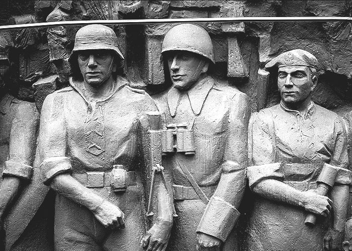 Czarno-białe zdjęcie: Figury trzech marmurowych żołnierzy stoją obok siebie przodem do obiektywu. Dwóch z nich ma kaski, jeden z nich czapkę. 