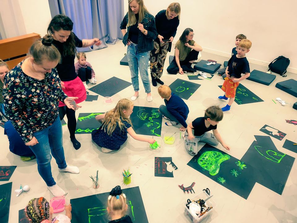 Kolorowe zdjęcie. Duże, jasnooświetlone pomieszczenie. Dzieci siedzą na podłogach i uwalniają swoją kreatywność na swoich dużych kartkach papieru. Pracom przyglądają się rodzice.